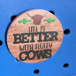 May include: A round wooden sign with a brown highland cow head and green leaves. The sign says "Life is Better with Fluffy Cows".
