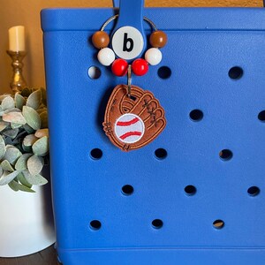 Puede incluir: Bolsa de mano azul con un amuleto de guante de b&eacute;isbol de cuero marr&oacute;n y un llavero de cuentas rojas y blancas con un amuleto de letra "b".