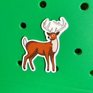 May include: A brown and white deer sticker with large antlers on a green background.