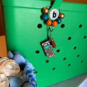 Pu&ograve; includere: Una borsa tote verde brillante con un ciondolo decorativo. Il ciondolo presenta la lettera "b" su sfondo bianco, circondata da perline colorate in oro, turchese e arancione. Un piccolo portachiavi con nappa e ciondolo decorativo &egrave; attaccato.