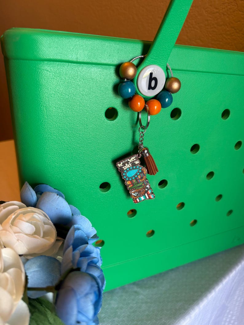 Pu&ograve; includere: Una borsa tote verde brillante con un ciondolo decorativo. Il ciondolo presenta la lettera "b" su sfondo bianco, circondata da perline colorate in oro, turchese e arancione. Un piccolo portachiavi con nappa e ciondolo decorativo &egrave; attaccato.