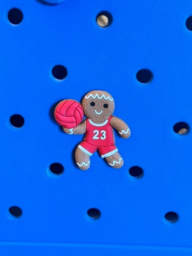 May include: A gingerbread man wearing a red jersey with the number 23 on it, holding a red basketball. The gingerbread man is brown with white icing details and a smiling face.