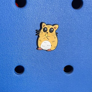 May include: A yellow cartoon hamster charm with black eyes and a pink nose. The charm is on a blue background with black holes.