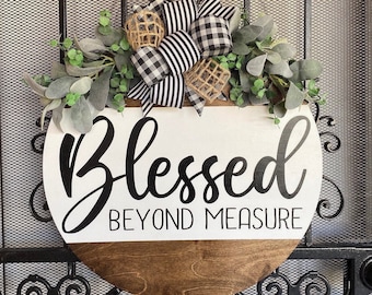 May include: A round wooden sign with the words "Blessed Beyond Measure" in black script. The top half is white, the bottom is stained brown. It's decorated with greenery, a black and white checkered bow, and burlap accents.