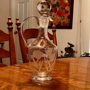 May include: A clear glass decanter with a bulbous stopper and a handle. The decanter features an etched floral design of tulips. The decanter is sitting on a wooden table.