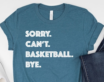 May include: Teal t-shirt with the text "SORRY. CAN'T. BASKETBALL. BYE." paired with dark blue jeans and white sneakers. The t-shirt is folded at the bottom. The sneakers are white canvas.