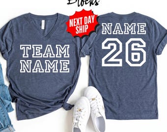 May include: A dark blue heathered V-neck t-shirt featuring the white text TEAM NAME on the front and the word NAME above the number 26 in white on the back. The shirt is displayed alongside a pair of blue jeans and white sneakers.
