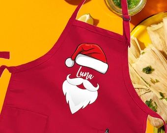 May include: A red apron with a Santa Claus design, the name "Luna" is printed on the apron. The apron is next to a plate of tamales, lemons, and a jar of green sauce. The word "Red" is printed on the bottom right.