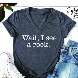 May include: A dark grey V-neck t-shirt with the text "Wait, I see a rock." printed in white on the front.