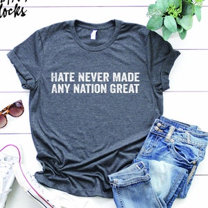 May include: A dark gray t-shirt with the text "Hate Never Made Any Nation Great" printed in white on the front. The shirt is laid flat on a white wooden surface with a pair of sunglasses, a pair of jeans, and a pair of white sneakers.