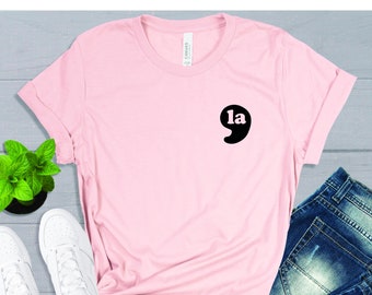 May include: Pink t-shirt with a black graphic of a comma with the word "la" inside. The shirt is laid flat on a gray surface with a pair of white sneakers and blue jeans.