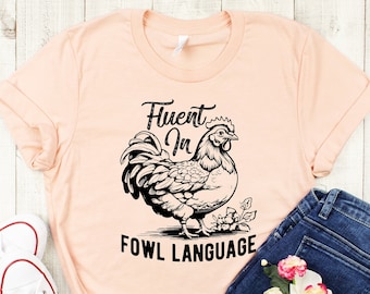 May include: A peach-colored t-shirt with a black graphic of a rooster and the text "Fluent in Fowl Language". The shirt is paired with blue jeans and white sneakers.
