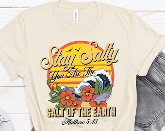 May include: A beige t-shirt with a retro-style graphic design featuring a sunset, waves, and flowers. The text on the shirt reads "Stay Salty You Are The Salt Of The Earth Matthew 5:13".