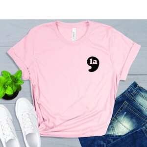 May include: Pink t-shirt with a black graphic of a comma with the word "la" inside. The shirt is laid flat on a gray surface with a pair of white sneakers and blue jeans.