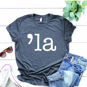 May include: A dark grey t-shirt with a white graphic of an apostrophe and the word "'la" printed on the front.