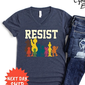 May include: Navy blue v-neck t-shirt with the word "RESIST" in a bold, yellow font. Below the text are colorful cartoon figures. The shirt is paired with blue jeans. The image also includes a red starburst graphic with the words "NEXT DAY SHIP".