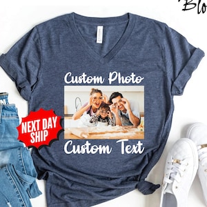 May include: A dark blue heathered V-neck t-shirt featuring a central rectangular graphic with the text Custom Photo above and Custom Text below. The graphic shows a family of three in a kitchen making dough, with the text NEXT DAY SHIP in red.