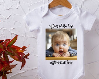 May include: White baby onesie with a wooden hanger. The onesie features a photo of a baby with the text "custom photo here" above the photo and "custom text here" below it. A vase of red and yellow leaves is to the left.
