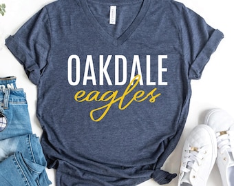 May include: A navy blue V-neck t-shirt with the words "OAKDALE eagles" in white and yellow script. The shirt is displayed with a pair of white sneakers, blue jeans, and sunglasses.