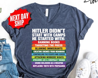 May include: A heather navy blue V-neck t-shirt featuring white text that reads: HITLER DIDN'T START WITH CAMPS HE STARTED WITH: followed by a list of phrases in rainbow-colored bars including banning books, targeting the press, and blaming immigrants.