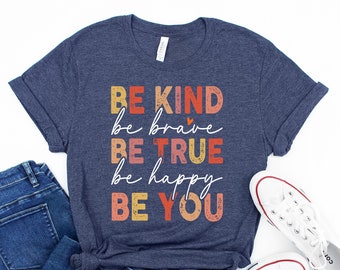 May include: A navy blue t-shirt with a vintage-style graphic that reads "Be Kind, Be Brave, Be True, Be Happy, Be You". The graphic is in shades of red, orange, yellow, and pink.