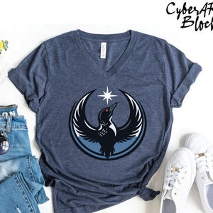 May include: Navy blue V-neck t-shirt featuring a graphic of a bird with outstretched wings, a star, and the text "CyberART Blocks". The bird is black with white and blue accents. The shirt is paired with blue jeans and white sneakers.