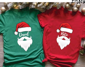 May include: Two short-sleeved t-shirts, one green and one red, each featuring a Santa Claus face design. The design includes a red hat, white beard, and the names "David" and "Katie" in white script. The shirts are likely for Christmas.