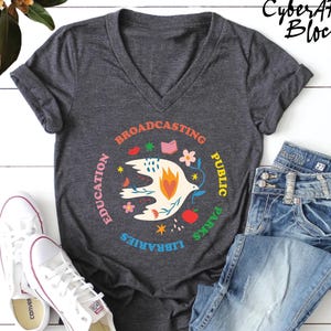 May include: A dark gray V-neck t-shirt featuring a central graphic of a white bird with a flame heart, surrounded by the text Education, Broadcasting, Public, Parks, and Libraries, accompanied by small colorful icons of flowers, a book, and an apple.
