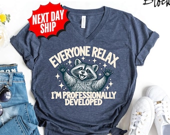May include: A heather navy blue V-neck t-shirt featuring a graphic of a raccoon with its arms raised, surrounded by small stars. The text reads: EVERYONE RELAX I'M PROFESSIONALLY DEVELOPED. The shirt is displayed with blue jeans, sunglasses, and white sneakers.