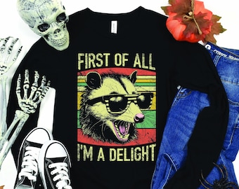 May include: Black long-sleeve shirt with a graphic of an opossum wearing sunglasses, with the text "FIRST OF ALL I'M A DELIGHT". The design has a vintage, distressed look. The shirt is paired with blue jeans and black sneakers.