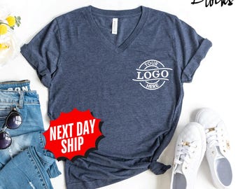 May include: A heathered navy blue V-neck t-shirt featuring a white circular graphic on the left chest with the text "YOUR LOGO HERE" inside. The shirt is displayed alongside blue denim jeans, white sneakers, and black sunglasses.