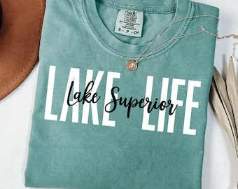 May include: A sage green short-sleeve t-shirt featuring the white text "LAKE LIFE" in a bold sans-serif font with the black cursive text "Lake Superior" layered over it. The shirt has a Comfort Colors brand label at the inner neckline.