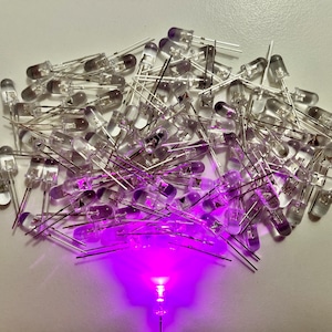 Könnte beinhalten: Eine große Gruppe von klaren LED-Leuchten mit silbernen Leitungen, die in einem lockeren Haufen angeordnet sind. Eine der LEDs leuchtet und strahlt ein helles violettes Licht aus.