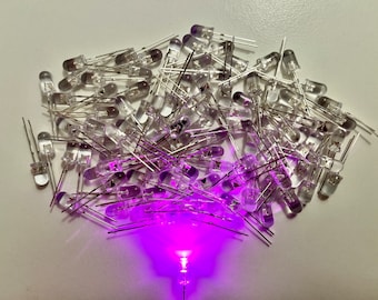 Puede incluir: Un gran grupo de luces LED transparentes con cables plateados, dispuestos en una pila suelta. Una de las LED está encendida y emite una luz púrpura brillante.