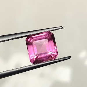 May include: A square-cut, vibrant pink gemstone held by a pair of dark metal tweezers. The gemstone features a faceted surface that reflects light, showcasing a clear, transparent quality with deep magenta hues throughout the stone's structure.