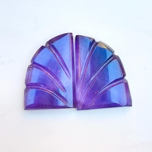 May include: Two matching, fan-shaped gemstones in shades of purple and blue. The gemstones are carved with parallel lines, creating a textured effect. The overall shape is a half-circle, with a flat base and a rounded top. The gemstones are likely amethyst.