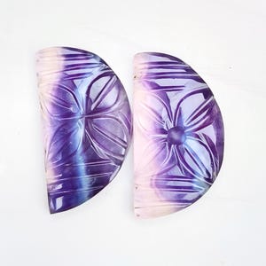 Könnte beinhalten: Zwei geschnitzte, halbmondförmige Fluorit-Steine. Die Steine sind eine Mischung aus Lila, Rosa und Weiß.