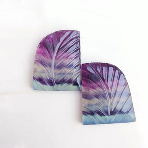 Könnte beinhalten: Zwei geschnitzte Fluorit-Cabochons mit einem Blattdesign. Die Cabochons sind eine Mischung aus Lila, Blau und Grün.