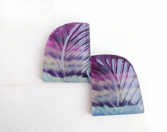 Puede incluir: Dos cabujones de fluorita tallados con un diseño de hoja. Los cabujones son una mezcla de morado, azul y verde.