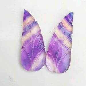 Könnte beinhalten: Zwei geschnitzte, blattförmige Cabochons aus Fluorit in Lila und Weiß. Die Cabochons sind durchscheinend und haben ein natürliches, organisches Aussehen.