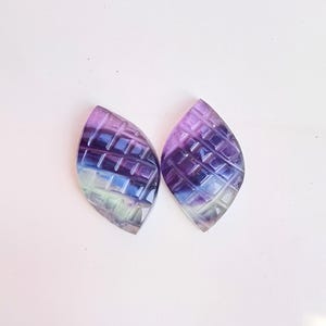 Könnte beinhalten: Zwei regenbogenfarbene Fluorite-Cabochons in Lila, Blau und Grün mit geometrischem Muster. Die Cabochons sind blattförmig und haben eine glatte, polierte Oberfläche.