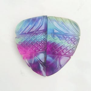 Könnte beinhalten: Ein geschnitzter Edelstein in schildförmiger Form, geteilt in zwei Hälften. Der Stein zeigt einen Farbverlauf von Blau, Lila, Rosa und Grün. Die Oberfläche weist aufwendige Schnitzmuster auf, die ein strukturiertes Aussehen erzeugen. Der Edelstein wird wahrscheinlich für Schmuck oder Dekorationszwecke verwendet.