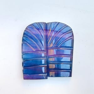 May include: A blue and purple iridescent carved stone with a geometric pattern. The stone is shaped like a leaf and has a smooth, polished surface.