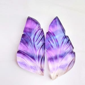 Könnte beinhalten: Zwei geschnitzte, blattförmige Cabochons aus Fluorit in den Farben Lila, Blau und Weiß. Die Cabochons sind durchscheinend und haben ein gestreiftes Muster.