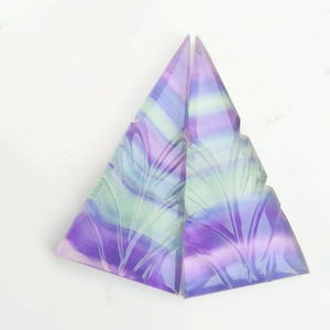 Könnte beinhalten: Zwei dreieckige Stücke geschnitzter Fluorit, eines lila und grün, das andere grün und rosa, vor weißem Hintergrund.