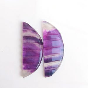 Könnte beinhalten: Zwei halbmondförmige, violette und weiße Fluorit-Cabochons. Die Cabochons haben ein gestreiftes Muster und sind durchscheinend.