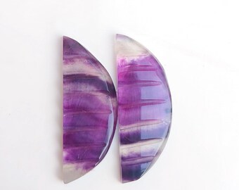 Puede incluir: Dos cabujones de fluorita en forma de media luna, de color morado y blanco. Los cabujones tienen un patrón estriado y son translúcidos.