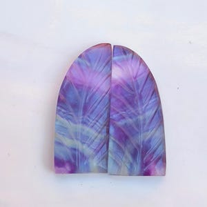 Könnte beinhalten: Zwei geschnitzte Fluorit-Cabochons mit einem lila, blauen und weißen Farbmuster. Die Cabochons haben die Form eines abgerundeten Rechtecks.