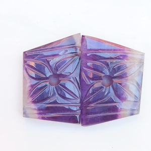 May include: Two carved amethyst gemstone cabochons with a floral design. The cabochons are a light purple color with a white center and are cut in a rectangular shape.