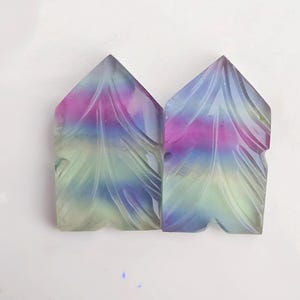 Könnte beinhalten: Zwei geschnitzte Fluorit-Steine mit einem Federdesign. Die Steine sind eine Mischung aus Grün, Blau, Lila und Rosa.